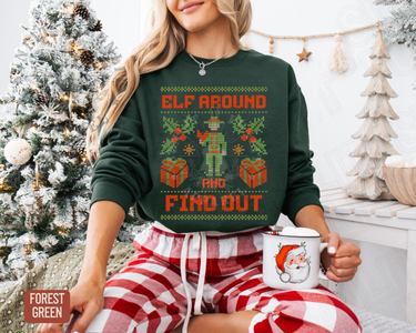 A woman wearing a green sweater with a Christmas-themed design is sitting in front of a Christmas tree, holding a mug with a Santa Claus image.